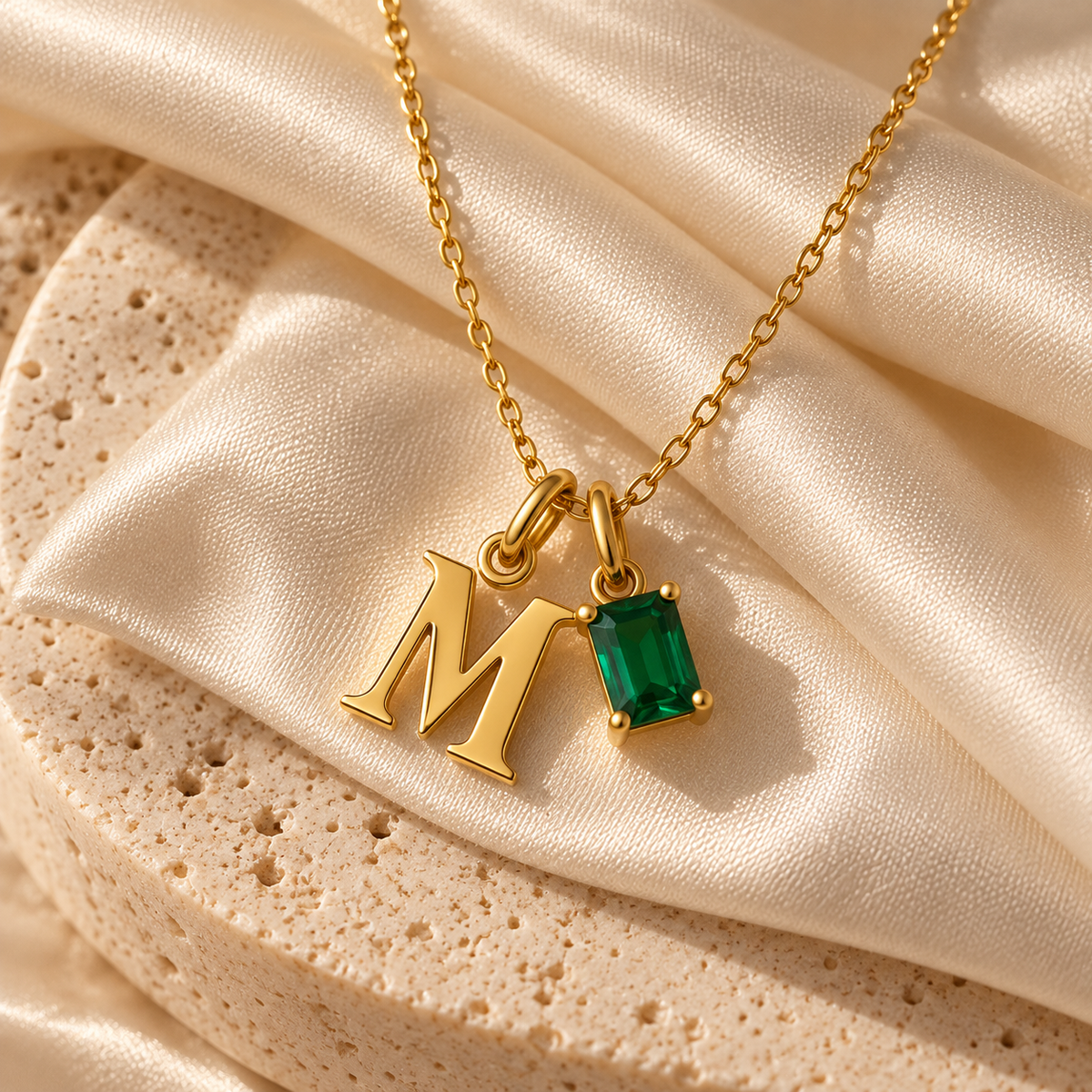 Gold initial necklace with a letter 'M' pendant and a rectangular green gemstone charm, displayed on draped champagne satin fabric.