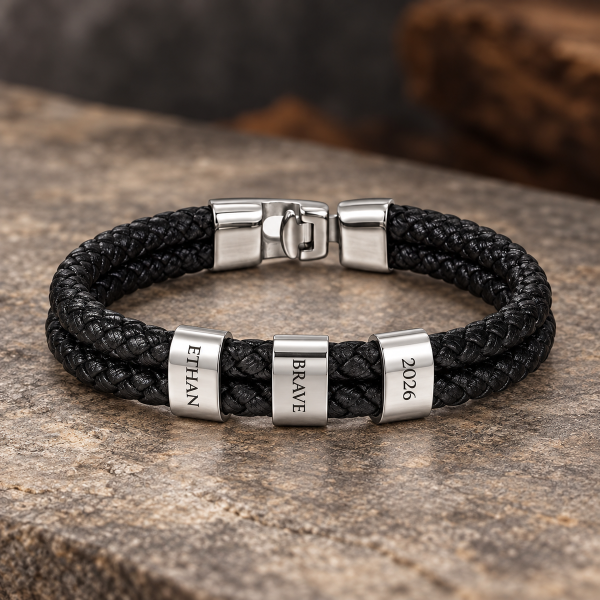 Personalized black braided leather bracelet with three silver engraved rings reading Ethan, Brave, and 2026.
