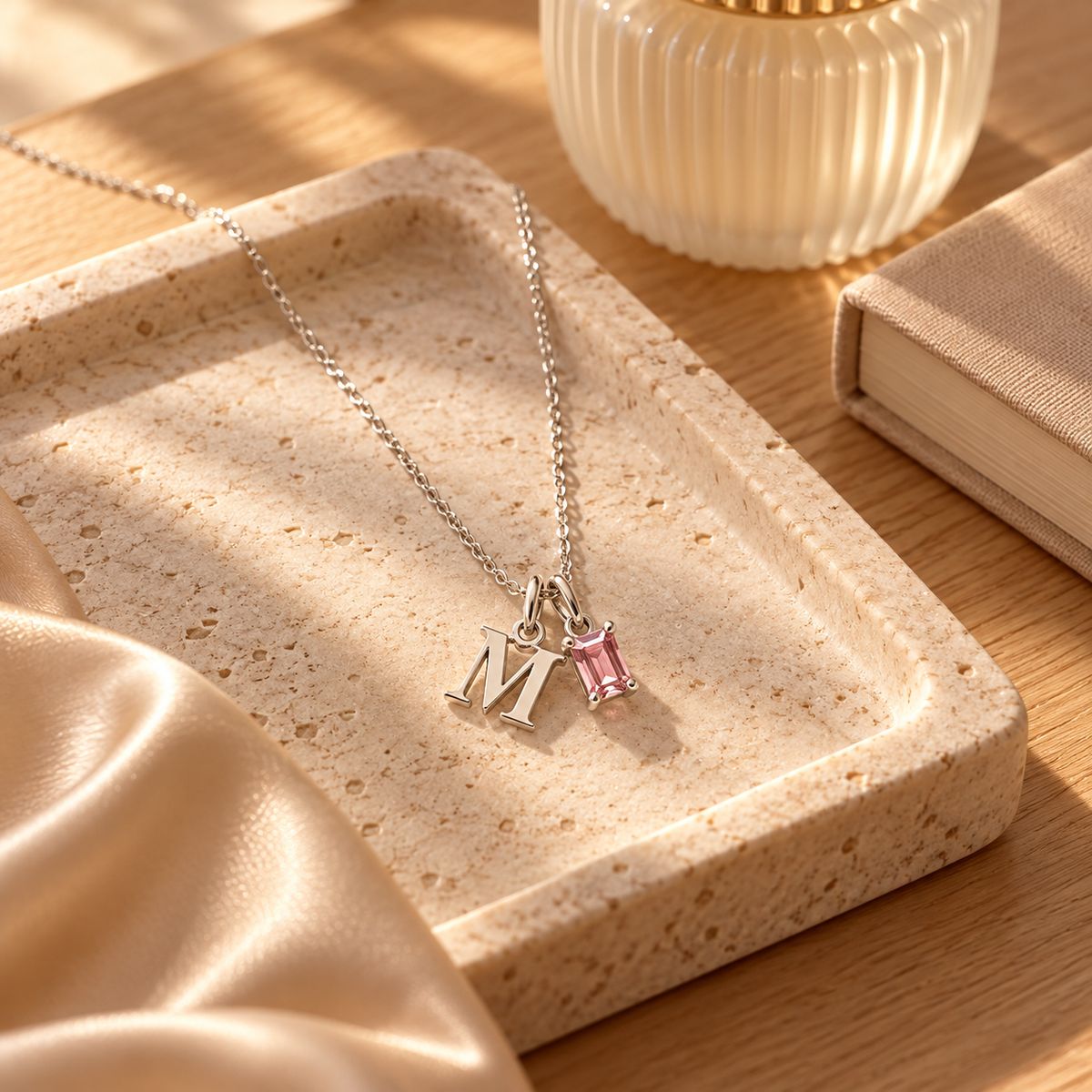 A silver initial necklace with a letter 'M' pendant and a small pink gemstone charm, displayed on a beige stone tray on a wooden table.