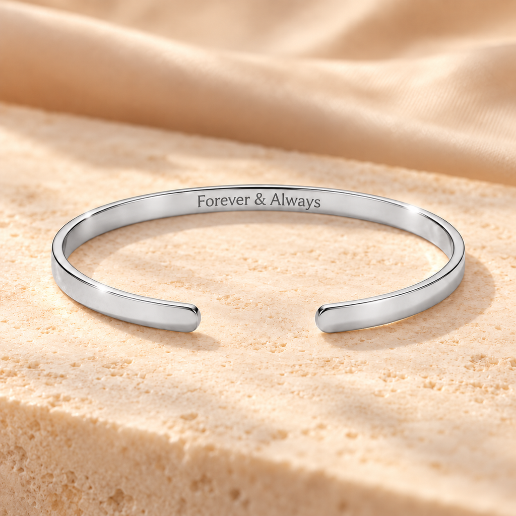 A polished silver open cuff bracelet engraved with the words 'Forever & Always', resting on a textured beige stone slab with draped beige fabric in the background.