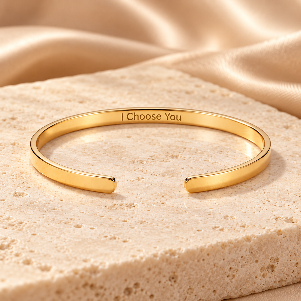 A polished gold open cuff bracelet engraved with the words 'I Choose You', displayed on a textured beige stone slab with draped satin fabric in the background.