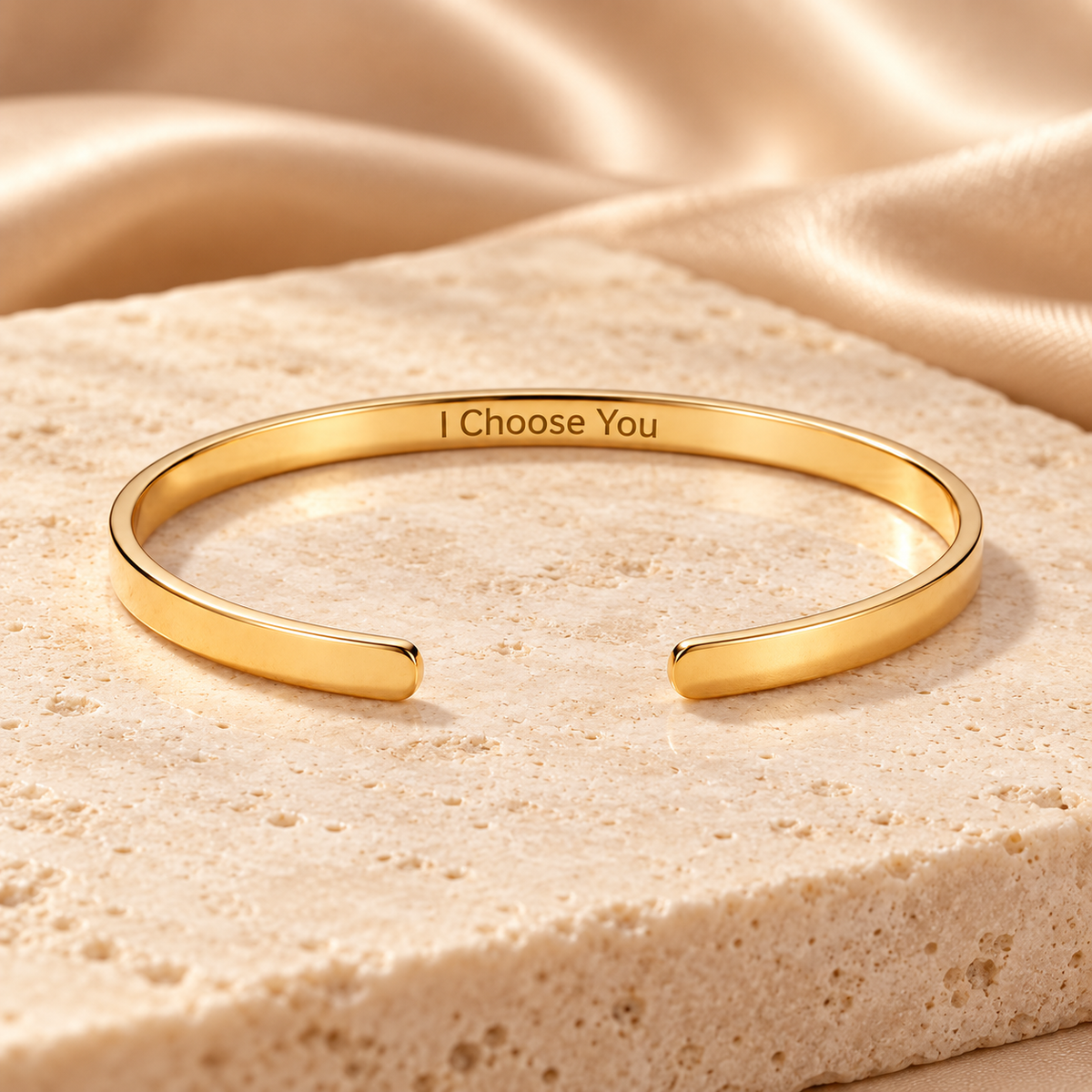 A polished gold open cuff bracelet engraved with the words 'I Choose You', displayed on a textured beige stone slab with draped satin fabric in the background.