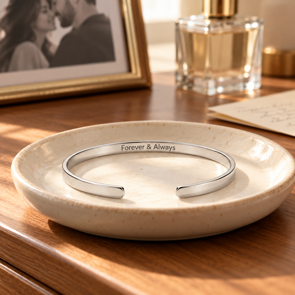 A polished silver cuff bracelet engraved with 'Forever & Always' sits in a shallow beige ceramic dish on a wooden table, with a blurred perfume bottle and framed photo in the background.