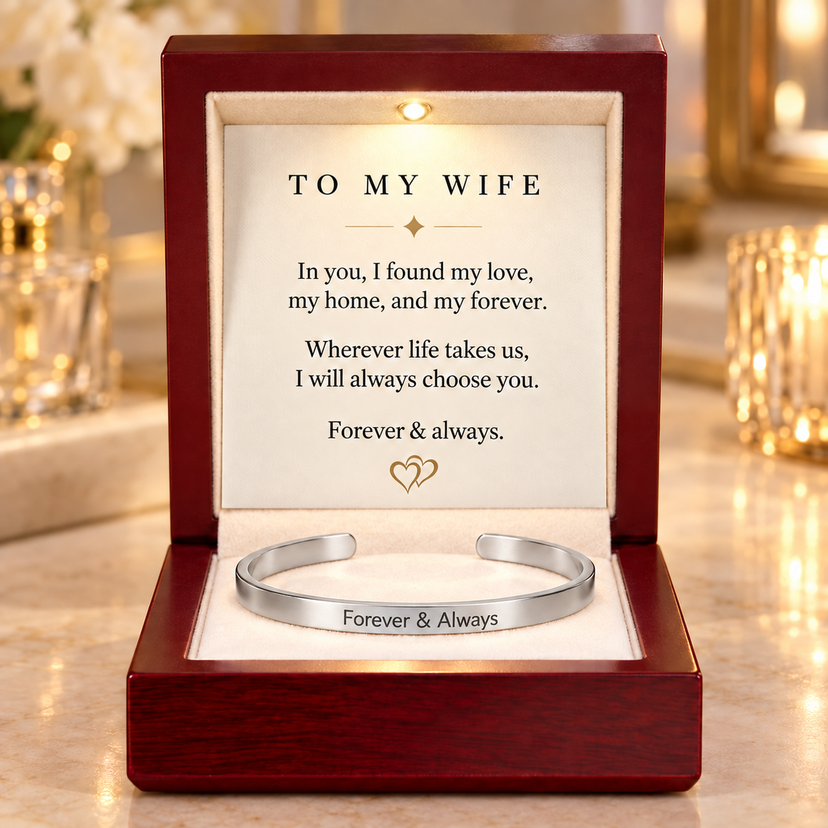 A silver open cuff bracelet engraved with 'Forever & Always' displayed inside a dark red wooden gift box. The inside lid features a cream-colored insert with a sentimental message addressed 'TO MY WIFE' and a small gold heart motif, set against a softly blurred warm background.